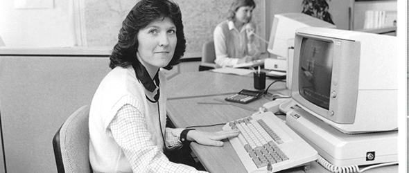 Black and white photo of a woman sat at a desk in an office with a computer